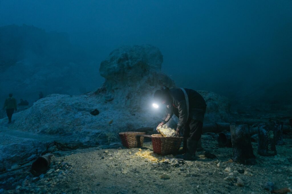 A solitary miner with headlamp gathers sulfur in baskets at a quarry during night.